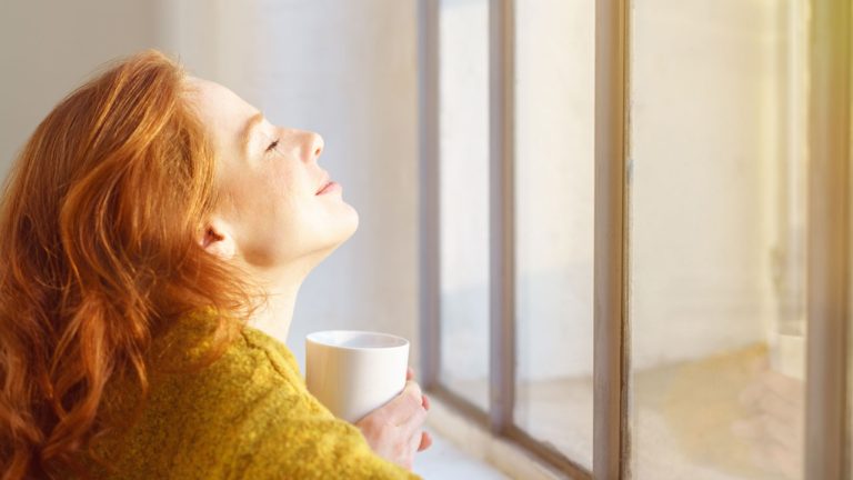 Mujer con una taza en la mano, recibiendo sol en su cara a través de una ventana - Vitamina D